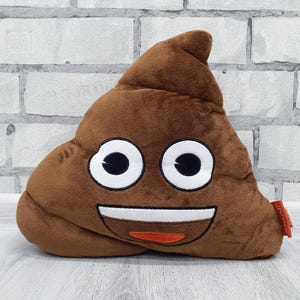 Op de afbeelding: Een bruin pluche emoji-kussen in de vorm van een lachende hoop poep. Het kussen heeft witte ogen, een witte glimlach en een oranje tong. Het kussen ligt op een lichtgrijze ondergrond voor een witte bakstenen muur.