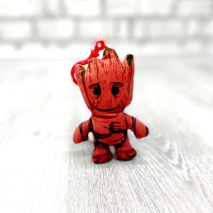 May include: A small, brown plush toy of a character from the Guardians of the Galaxy movie franchise. The toy is wearing a red shirt and has a green leafy head. The toy is standing on a white wooden surface.