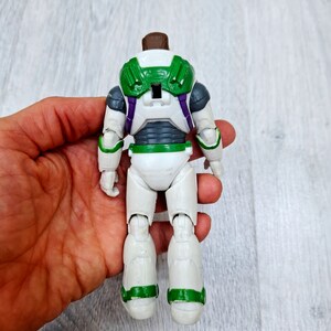 Puede incluir: Una figura de acci&oacute;n de Buzz Lightyear blanca y verde con una pieza de pecho morada y gris. La figura est&aacute; de pie sobre una superficie de madera.