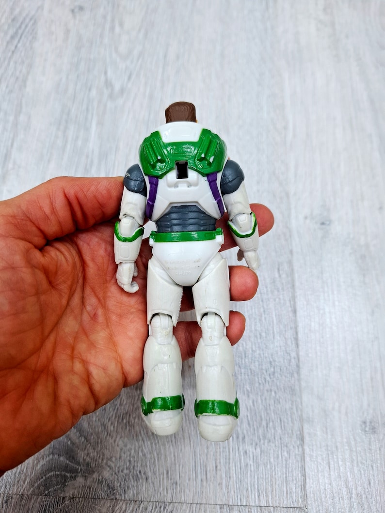 Puede incluir: Una figura de acci&oacute;n de Buzz Lightyear blanca y verde con una pieza de pecho morada y gris. La figura est&aacute; de pie sobre una superficie de madera.