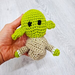 May include: A crocheted green and brown Yoda doll with black eyes. The doll is being held in a hand.