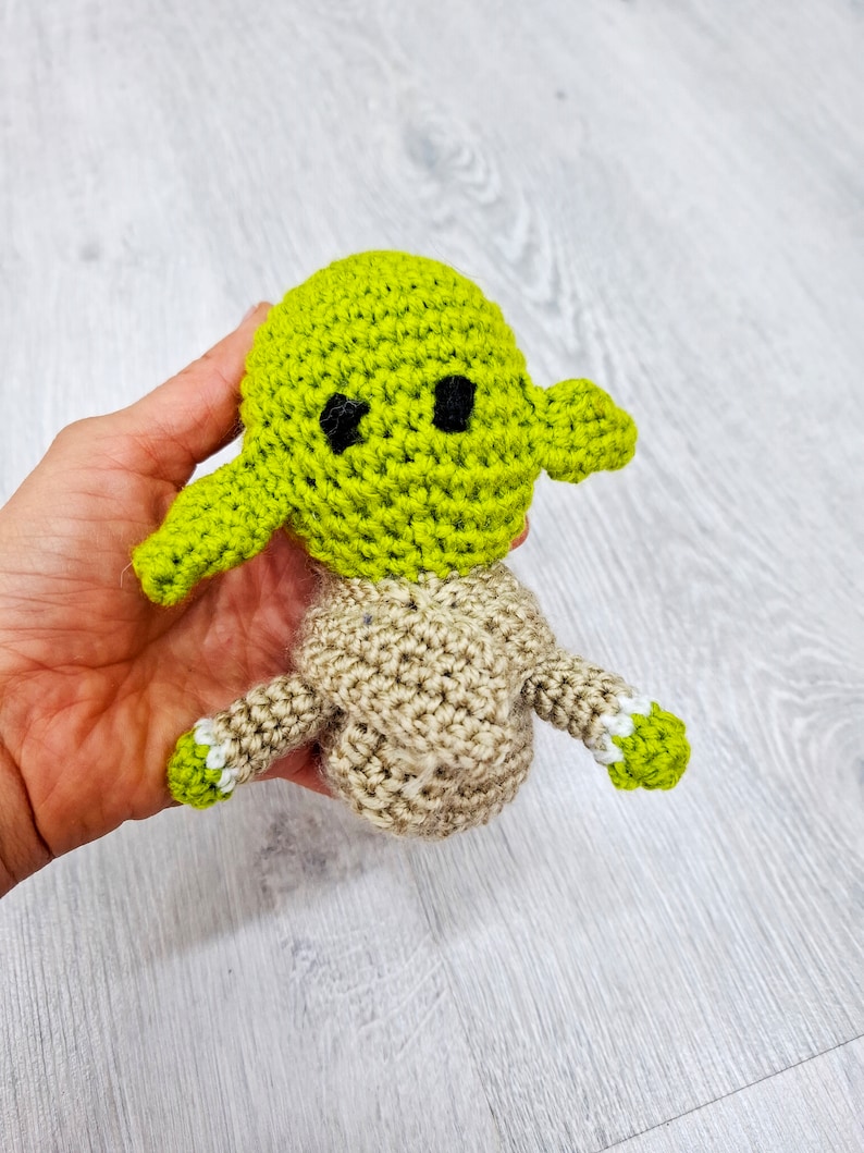 May include: A crocheted green and brown Yoda doll with black eyes. The doll is being held in a hand.