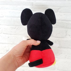 May include: A black and red plush Mickey Mouse toy with large ears. The toy is sitting on a white brick background.