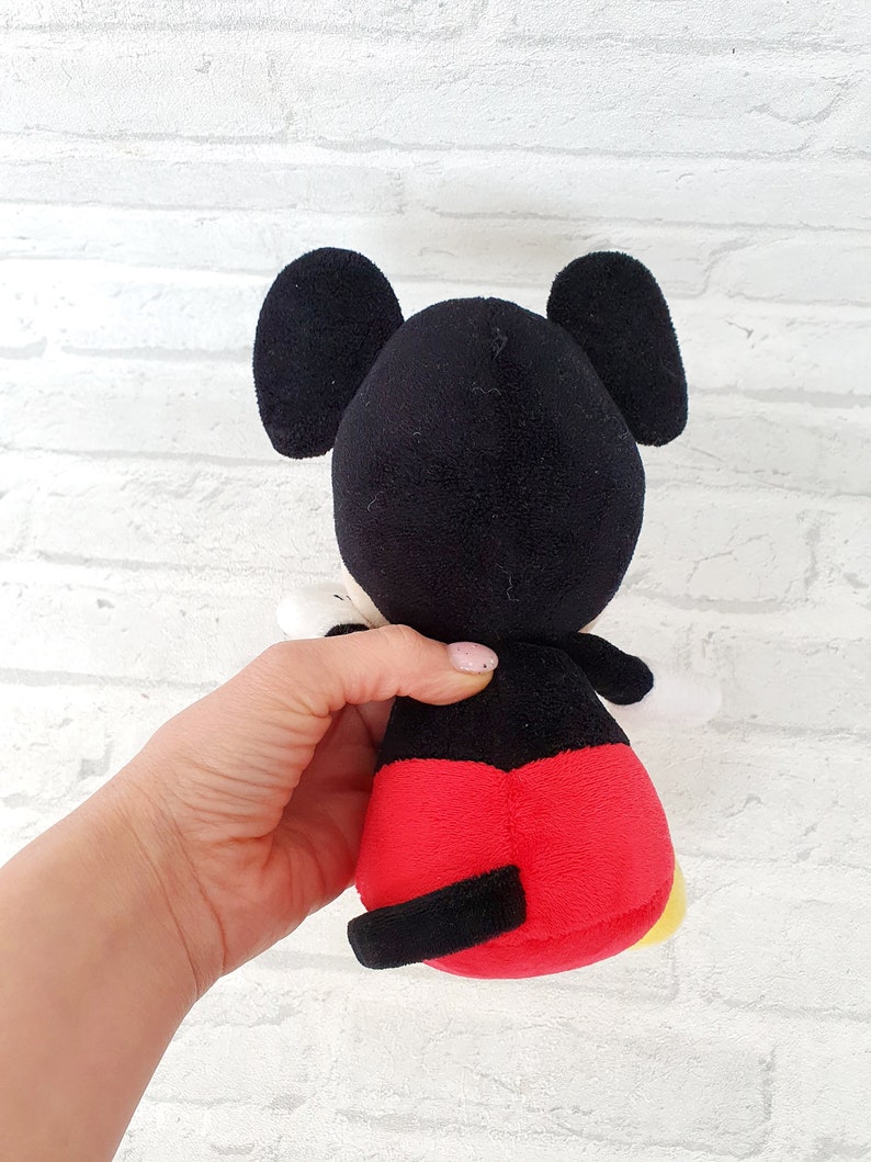 May include: A black and red plush Mickey Mouse toy with large ears. The toy is sitting on a white brick background.