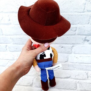 May include: A plush toy of Woody from the Toy Story franchise. Woody is wearing his signature brown cowboy hat, a yellow plaid shirt, a brown belt, blue jeans, and brown boots. The toy has a white tag on the back that says "Disney Pixar".