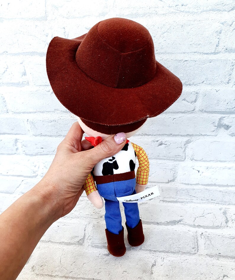May include: A plush toy of Woody from the Toy Story franchise. Woody is wearing his signature brown cowboy hat, a yellow plaid shirt, a brown belt, blue jeans, and brown boots. The toy has a white tag on the back that says "Disney Pixar".