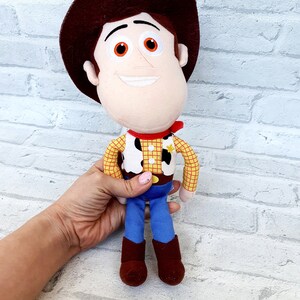 May include: A plush toy of Woody from the Toy Story franchise. Woody is wearing his signature brown cowboy hat, yellow and red plaid shirt, brown belt, blue jeans, and brown boots. He has a friendly smile and orange eyes.