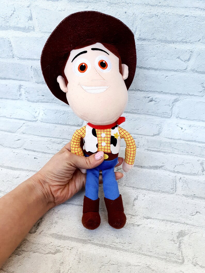 May include: A plush toy of Woody from the Toy Story franchise. Woody is wearing his signature brown cowboy hat, yellow and red plaid shirt, brown belt, blue jeans, and brown boots. He has a friendly smile and orange eyes.