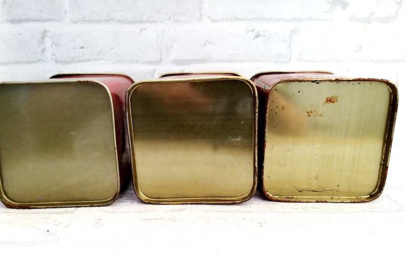 May include: Three vintage red metal canisters with gold lids. The canisters are empty and have a worn, vintage look.