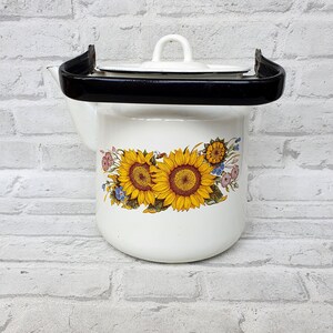 Könnte beinhalten: Weiße Email-Teekanne mit schwarzem Henkel und Deckel. Die Teekanne hat ein Blumenmuster mit Sonnenblumen und anderen Blumen in Gelb-, Blau- und Rosatönen.