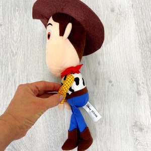 May include: Brown plush toy of Woody from Toy Story, wearing his signature cowboy hat, vest, and blue jeans. The toy has a white tag that reads "Disney PIXAR".