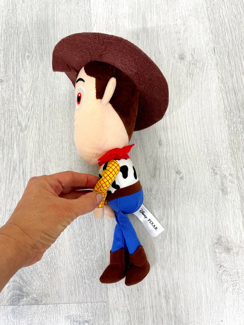 May include: Brown plush toy of Woody from Toy Story, wearing his signature cowboy hat, vest, and blue jeans. The toy has a white tag that reads "Disney PIXAR".