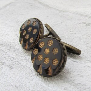 May include: A pair of cufflinks with a black background and gold floral pattern. The cufflinks are made of metal and have a round shape.