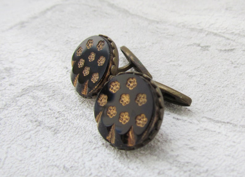 May include: A pair of cufflinks with a black background and gold floral pattern. The cufflinks are made of metal and have a round shape.