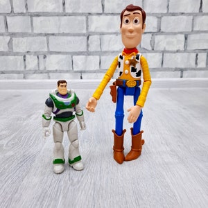 Puede incluir: Los personajes de Toy Story, Woody y Buzz Lightyear. Woody lleva su caracter&iacute;stico sombrero de vaquero y chaleco, mientras que Buzz est&aacute; en su traje de ranger espacial. Est&aacute;n de pie sobre un suelo de madera gris claro.