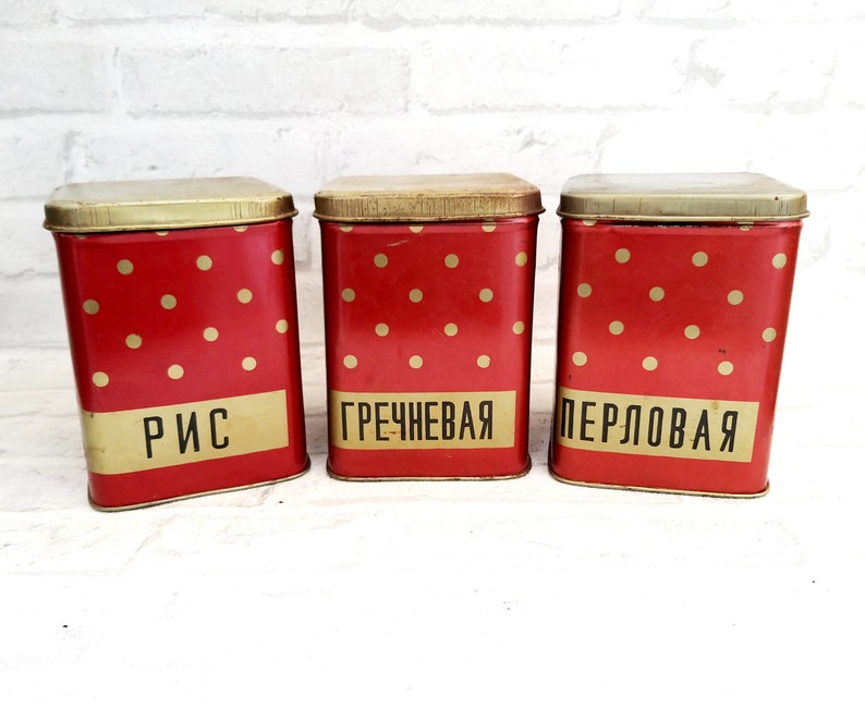 May include: Three red metal canisters with white polka dots and gold lids. The canisters are labeled "Rice", "Buckwheat", and "Pearl Barley" in black lettering.