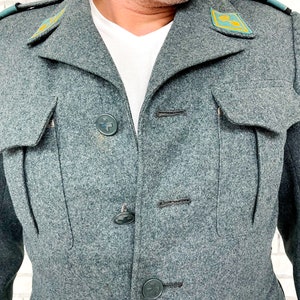 May include: A gray wool jacket with a high collar and four black buttons. The jacket has two flap pockets on the chest and two patch pockets on the bottom. There are two green embroidered patches on the shoulders.