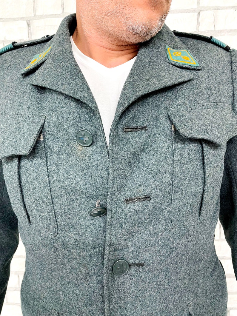 May include: A gray wool jacket with a high collar and four black buttons. The jacket has two flap pockets on the chest and two patch pockets on the bottom. There are two green embroidered patches on the shoulders.