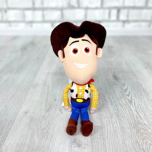 May include: A plush toy of Woody from Toy Story. He is wearing his signature brown cowboy hat, red bandana, yellow shirt with a brown vest, and blue jeans. He has a friendly smile and brown eyes.