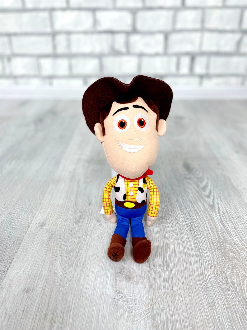 May include: A plush toy of Woody from Toy Story. He is wearing his signature brown cowboy hat, red bandana, yellow shirt with a brown vest, and blue jeans. He has a friendly smile and brown eyes.