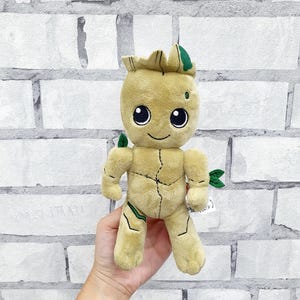 Puede incluir: Un peluche de un personaje con cuerpo marrón claro, ojos negros grandes y una cara sonriente. El juguete tiene detalles verdes en la cabeza y las extremidades, y se sostiene contra una pared de ladrillos blancos.