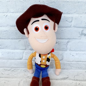 May include: A plush toy of Woody from the Toy Story franchise. Woody is wearing his signature brown hat, red and yellow plaid shirt, and blue jeans. He has a friendly smile and brown eyes.