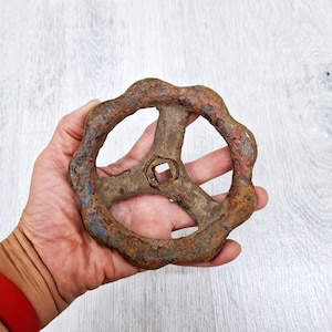 以下が含まれることがあります： 丸いハンドルと正方形の中央部を備えた、錆びたヴィンテージの金属製バルブホイール。ホイールは、赤と青の塗料の残骸があり、古さと使用感を示しています。インダストリアルデザインは、スチームパンクや素朴な装飾に最適です。