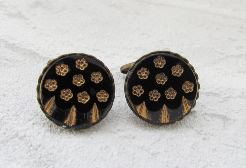 May include: A pair of black and gold cufflinks with a floral design. The cufflinks are round and have a gold rim.