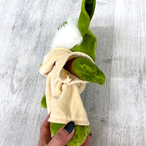 May include: Green plush toy with large ears and a white fur hood. The toy is wearing a tan robe and is being held by a hand.