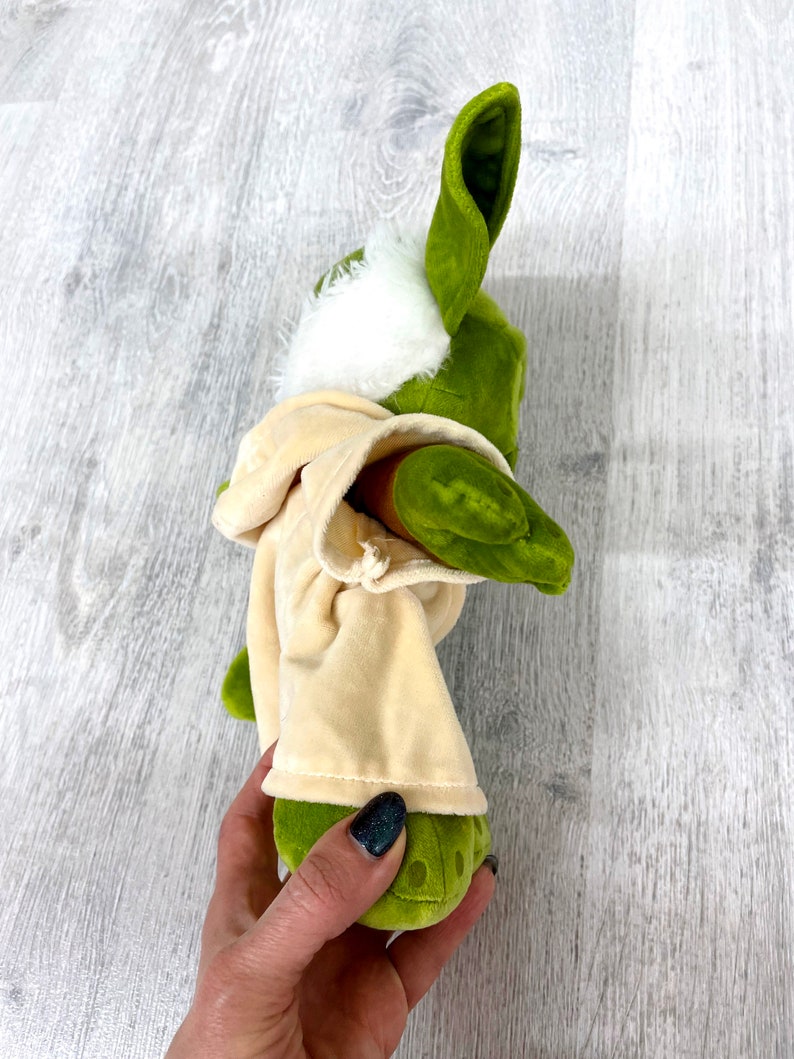 May include: Green plush toy with large ears and a white fur hood. The toy is wearing a tan robe and is being held by a hand.