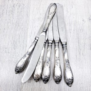 May include: A set of six antique silver butter knives. Each knife features a decorative, embossed handle with floral and scrollwork patterns. The blades are smooth and shiny. The knives are displayed on a light wooden surface.