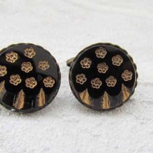 May include: A pair of cufflinks with a black background and gold floral pattern. The cufflinks are round and have a slightly raised edge.