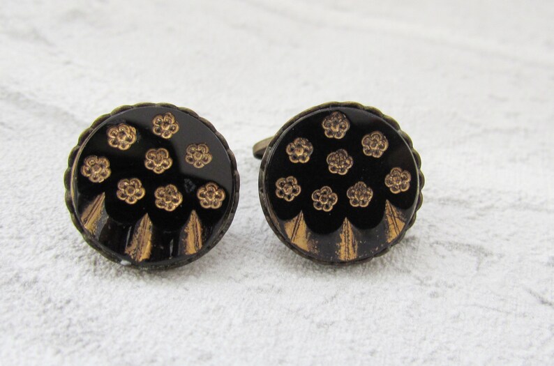 May include: A pair of cufflinks with a black background and gold floral pattern. The cufflinks are round and have a slightly raised edge.