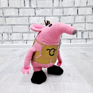 Puede incluir: Juguete de peluche rosa con forma de oso hormiguero con un chaleco marrón y la letra "C" en él. El juguete tiene zapatos negros y un mechón de pelo marrón en la cabeza.