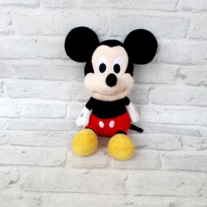 May include: A plush Mickey Mouse toy with black ears, a black nose, and red shorts with white buttons. The toy has yellow shoes and a white background.