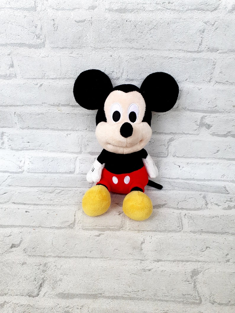May include: A plush Mickey Mouse toy with black ears, a black nose, and red shorts with white buttons. The toy has yellow shoes and a white background.