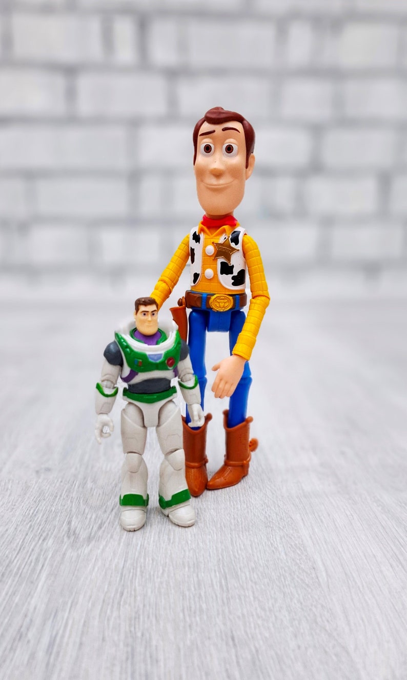 Puede incluir: Figuras de juguete de Woody y Buzz Lightyear de la franquicia Toy Story. Woody lleva su sombrero de vaquero, chaleco y pantalones vaqueros caracter&iacute;sticos. Buzz Lightyear lleva su traje de ranger espacial. Las figuras est&aacute;n de pie sobre una superficie de madera clara.