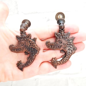 Könnte beinhalten: Zwei antike Schrankgriffe mit Drachenmotiv. Die Griffe sind aus Metall und haben eine dunkelbraune Oberfläche. Jeder Griff zeigt einen Drachenkopf mit Krone und einem eingerollten Schwanz.