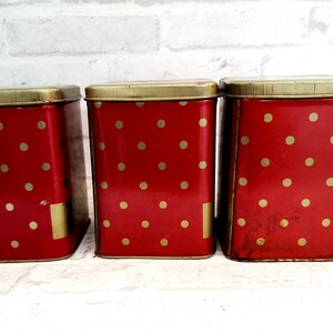 May include: Three red metal canisters with gold polka dots and gold lids. The canisters are lined up in a row.