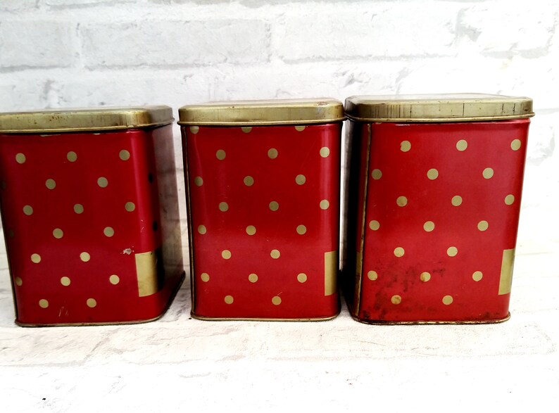 May include: Three red metal canisters with gold polka dots and gold lids. The canisters are lined up in a row.