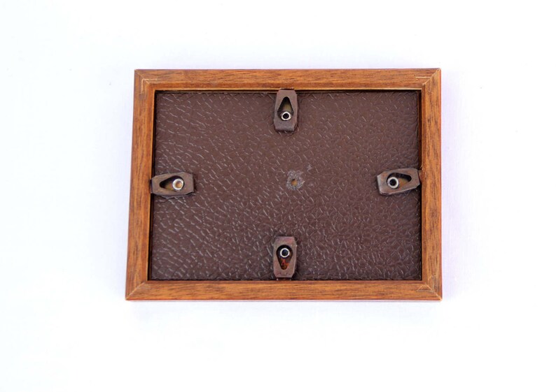 Puede incluir: Un marco de fotos rectangular de madera, visto desde atr&aacute;s, con un respaldo marr&oacute;n y herrajes met&aacute;licos para colgar. El marco est&aacute; hecho de madera de tonos c&aacute;lidos y tiene un dise&ntilde;o simple y cl&aacute;sico. La parte posterior tiene una apariencia texturizada similar al cuero.