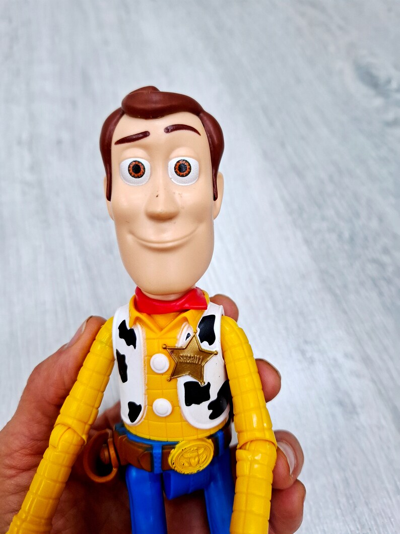 Puede incluir: Una figura de juguete de pl&aacute;stico amarillo y marr&oacute;n de Woody de la franquicia Toy Story. Woody lleva un pa&ntilde;uelo rojo, un chaleco amarillo con estampado de vaca blanco y negro, un cintur&oacute;n marr&oacute;n con hebilla dorada y pantalones vaqueros azules. Lleva una insignia de estrella de sheriff dorada en su chaleco que dice "Sheriff".