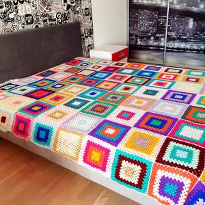 Peut inclure: Une couverture au crochet faite à la main avec un motif patchwork. La couverture présente une variété de carrés aux couleurs vives dans les tons de rose, bleu, jaune, vert et violet. La couverture est posée sur un lit, mettant en valeur son motif complexe et sa texture.