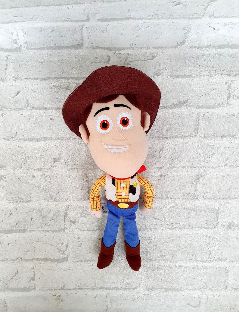 May include: A plush toy of Woody from Toy Story. He is wearing his signature brown hat, red bandana, yellow and brown plaid shirt, and blue jeans. He has a friendly smile and brown boots.