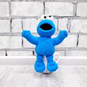 May include: A blue plush Cookie Monster toy sitting on a white ceramic mug. The toy has a big open mouth and large black eyes.