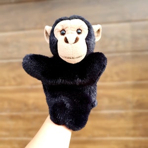 May include: A black plush monkey puppet with brown eyes and a brown nose. The puppet is being held up by a hand.