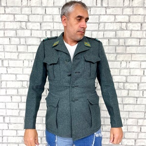 May include: A dark green military-style jacket with a button closure and two large pockets. The jacket has a collar and is made of a thick, textured fabric.