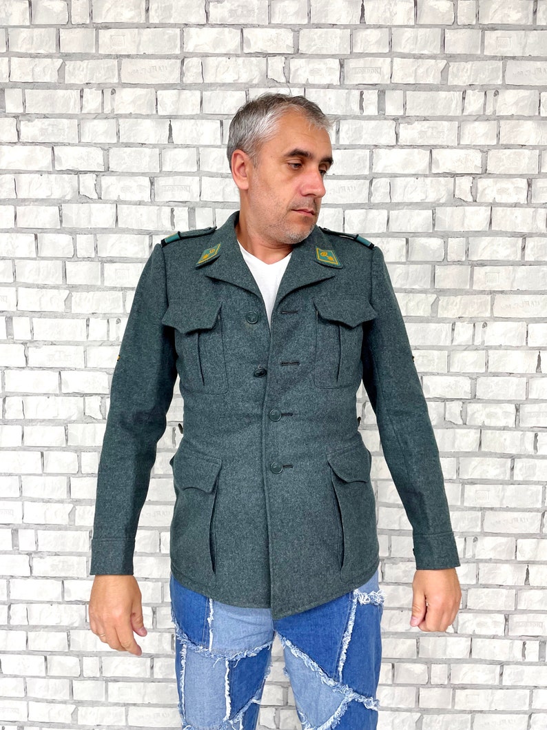May include: A dark green military-style jacket with a button closure and two large pockets. The jacket has a collar and is made of a thick, textured fabric.