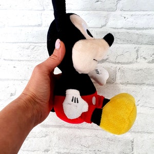 May include: A black and red plush Mickey Mouse toy with yellow shoes. The toy is sitting on a white brick wall.