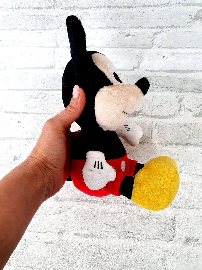 May include: A black and red plush Mickey Mouse toy with yellow shoes. The toy is sitting on a white brick wall.